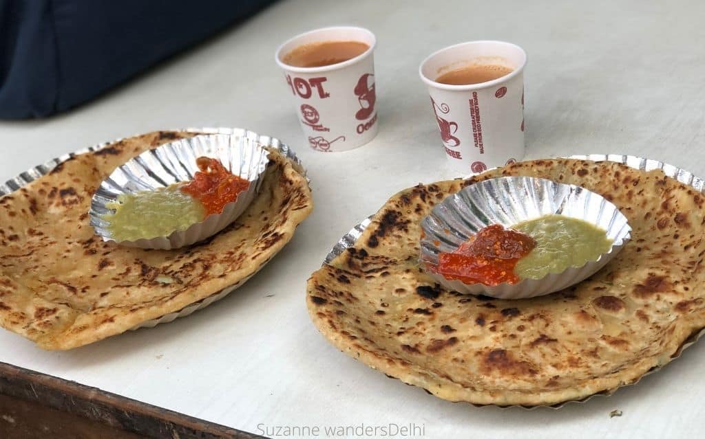 two plates of paranthas side by side with each with a smaller plate of assorted pickes on top and two paper cups of chai