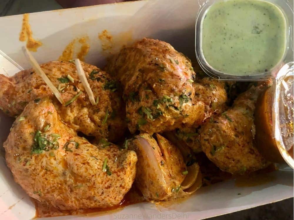 a serving on chicken tandoori momos smothered in creamy sauce with side of green chutney in paper container