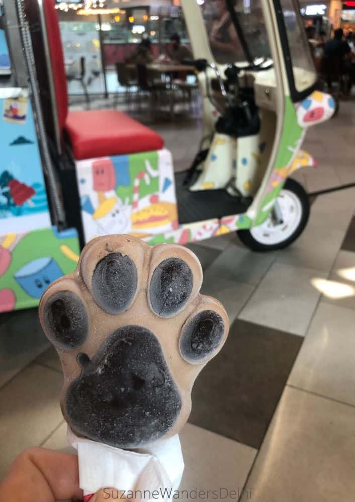 An ice cream coffee stick in the shape of a dog paw being held in front of the eMoi kiosk - definitely one of the best ice cream places in Delhi