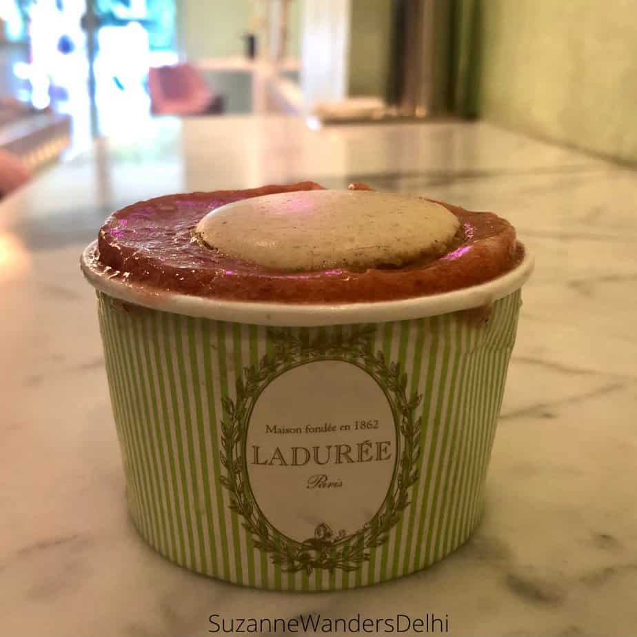 A Laduree cup filled to the brim with Raspberry sorbet and topped with a white macaron on a white marble counter top