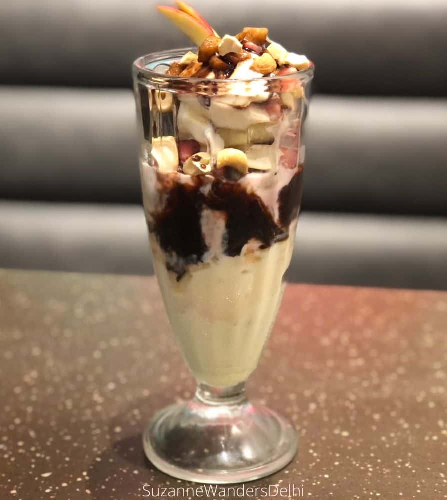 A tall, glass sundae dish filled with vanilla ice cream, chocolate sauce, nuts and fruits on a table