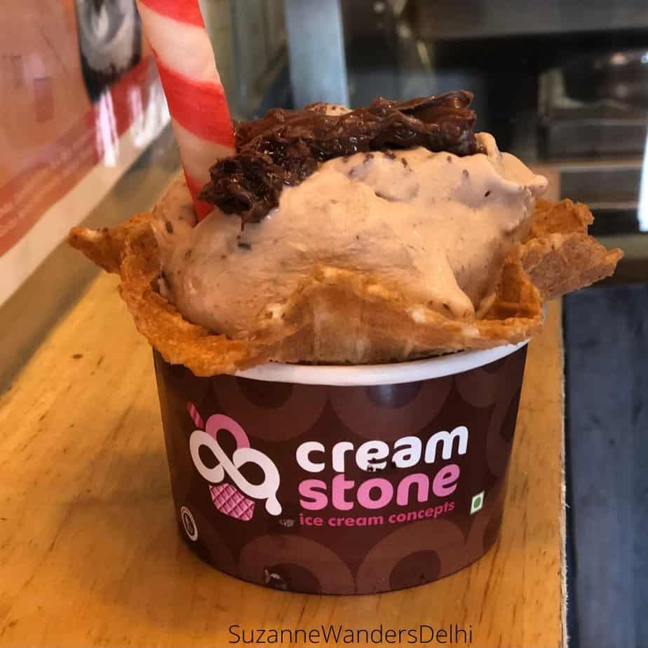 A scoop of Choco Strawberry ice cream in a waffle cup, topped with crumbled brownie and a pink and white twirly wafter cookie in a cream stone cup on a wooden counter