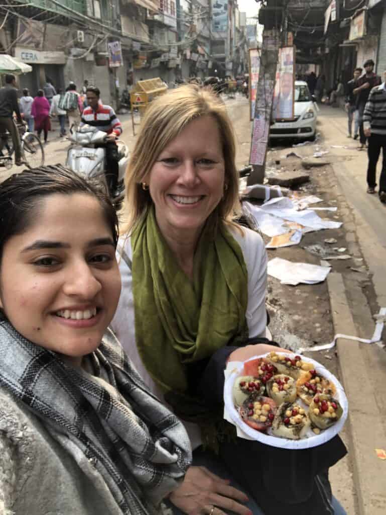 the author and a friend sitting outside on the a paper strewn street eating a plate of fruit chaat