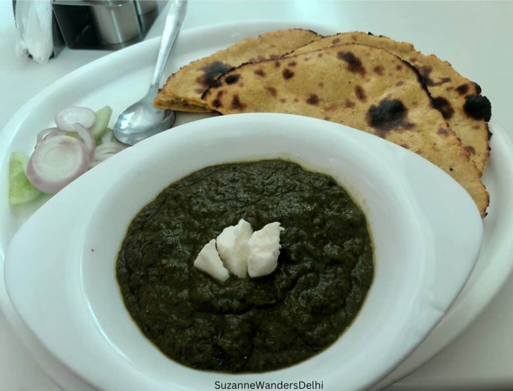 A white bowl and plate full of sarson ka saag with white butter on the top and makki ki rotis, with a side of sliced onion and cucumber