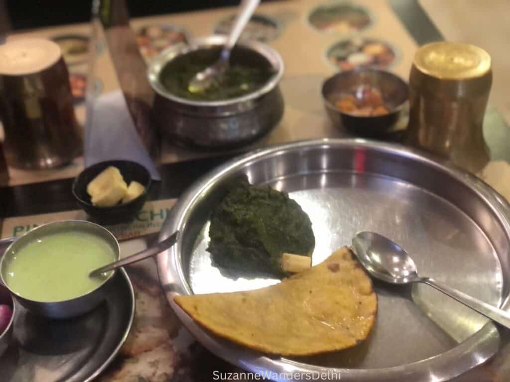 A round silver plate with sarson ka saag and half a roti on it - pots of chutney and sauces in the background