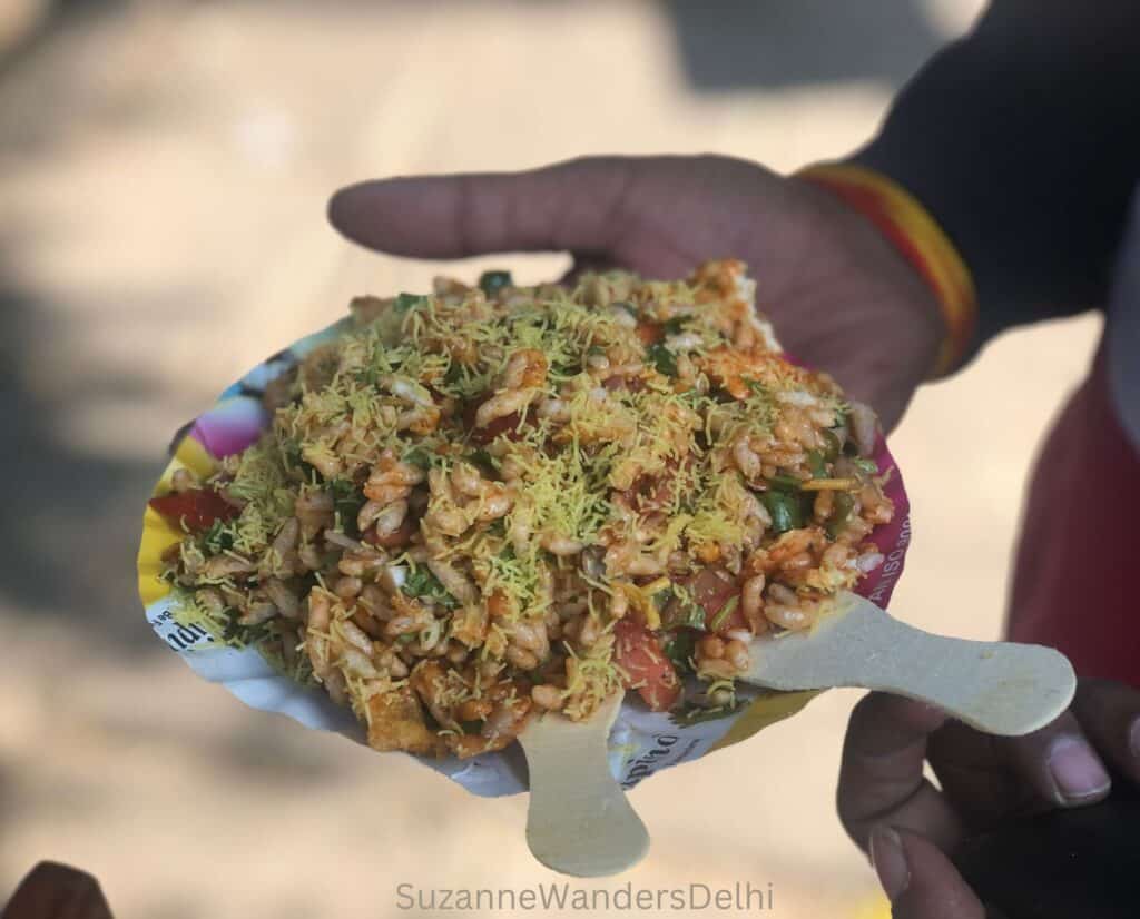 A plate of bhel puri being held in one hand with two wooden fork.  Bhel puri is one of the most popular Delhi street foods.