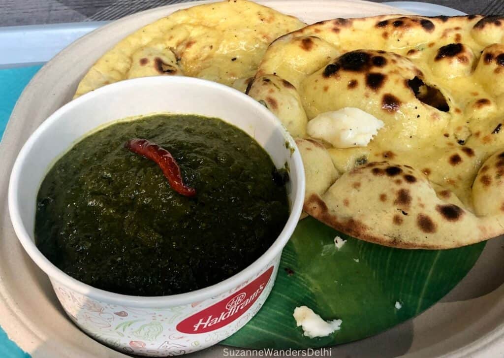 A bowl of sarson ka saag in a Haldiram's container on plate with 2 makki ki rotis