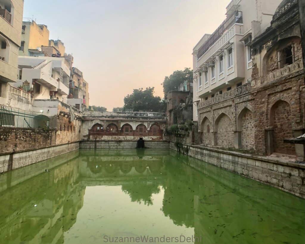 long view of the green water of the baoli, completely hemmed in by construction on 2 sides and an arched covered walkway at the far end