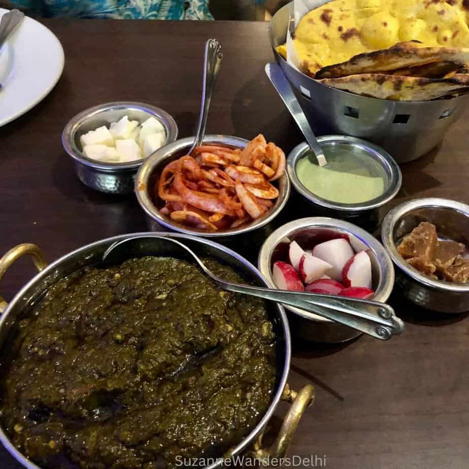 Silver serving dishes full of sarson ka saag, green chutney, white butter, jaggery, radishes, pickled onion and makki ki rotis at Gulati, one of the best places in Delhi for sarson ka saag and makki ki roti