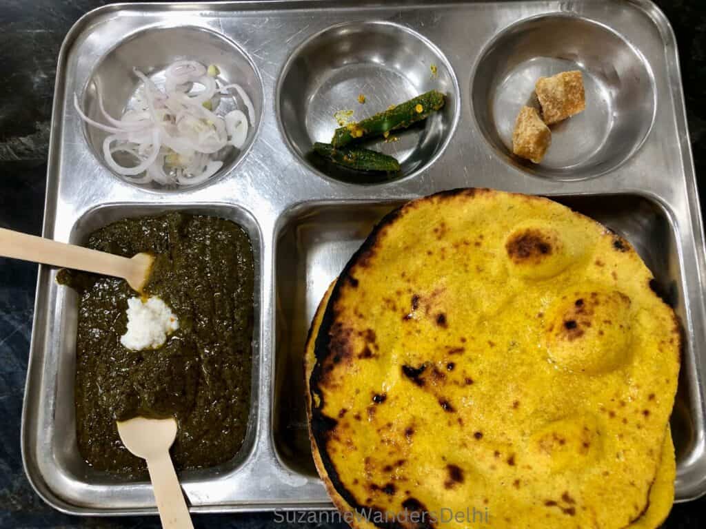 a metal thali tray filled with sliced onion, chilis, jaggery, sarson ka saag and tandoori baked makki ki roti at Dhania da Dhaba in Amar Colony, one of the best places for sarson ka saag and makki ki roti in Delhi