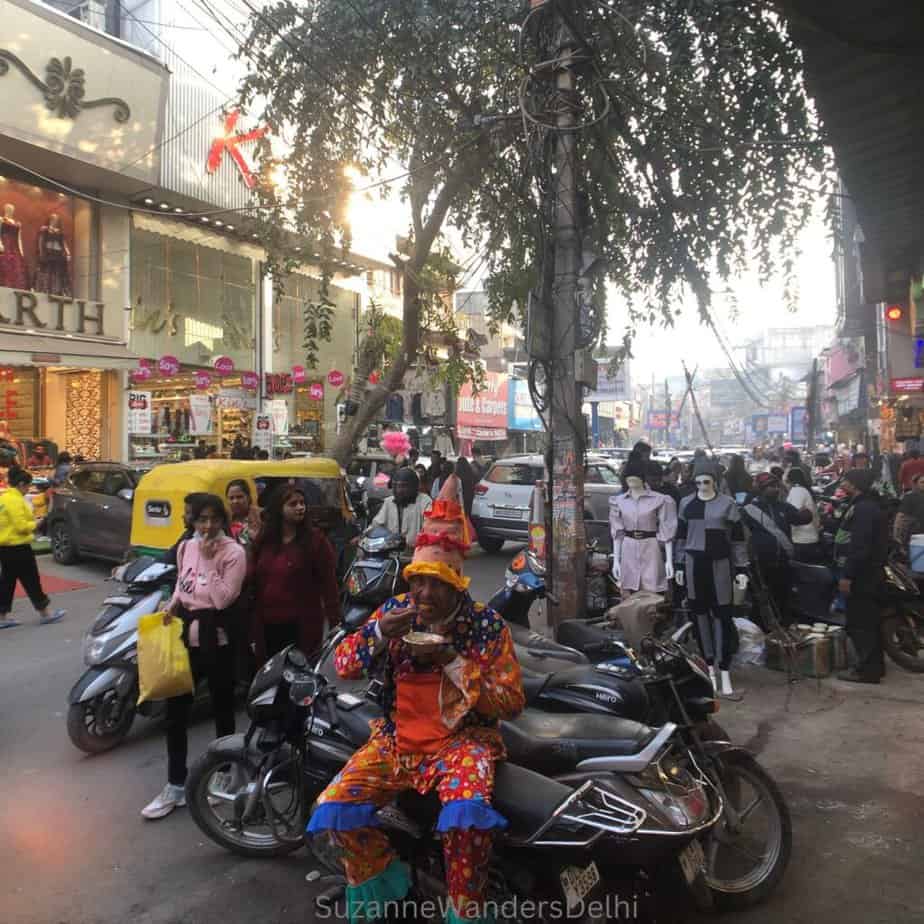 A clown sitting side saddle on a parked motorcycle eating a plate of chaat in the busy market