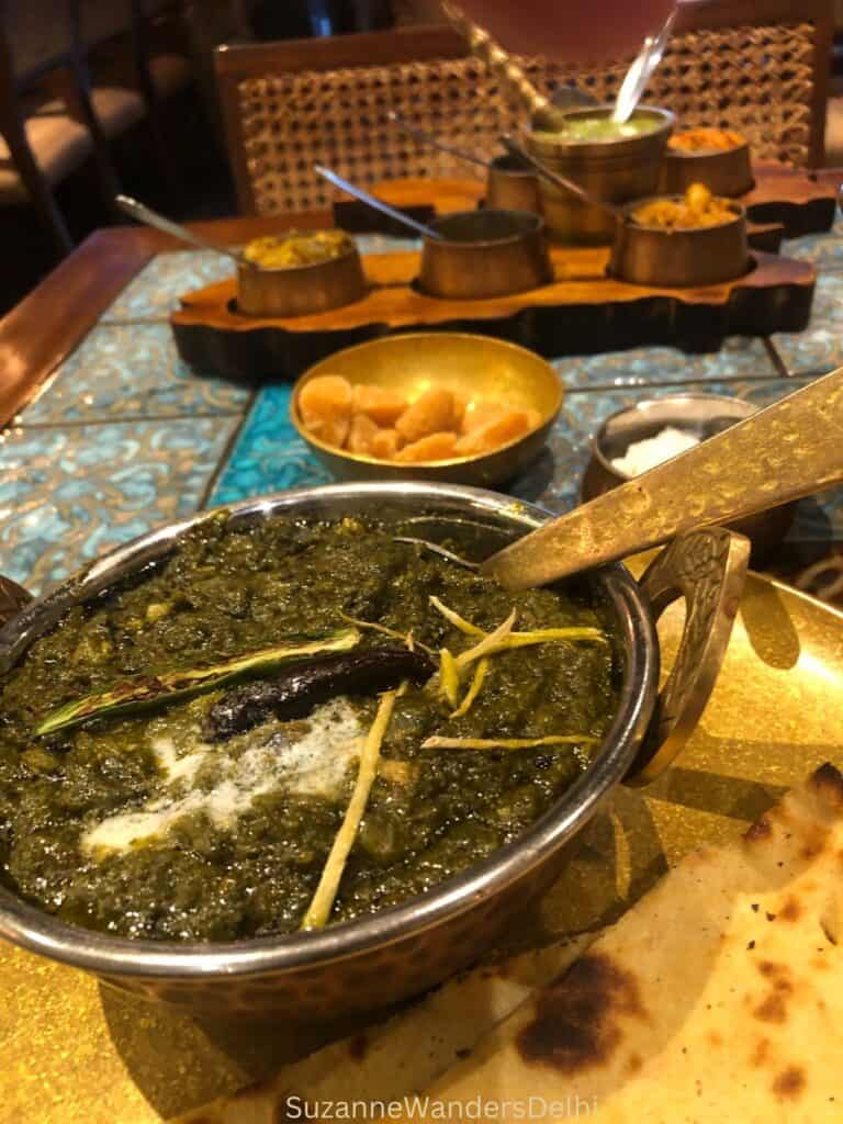 Big brass bowl of sarson ka saag on a gold plate with a roti on a tiled table.  Small pots of condiments in the background.