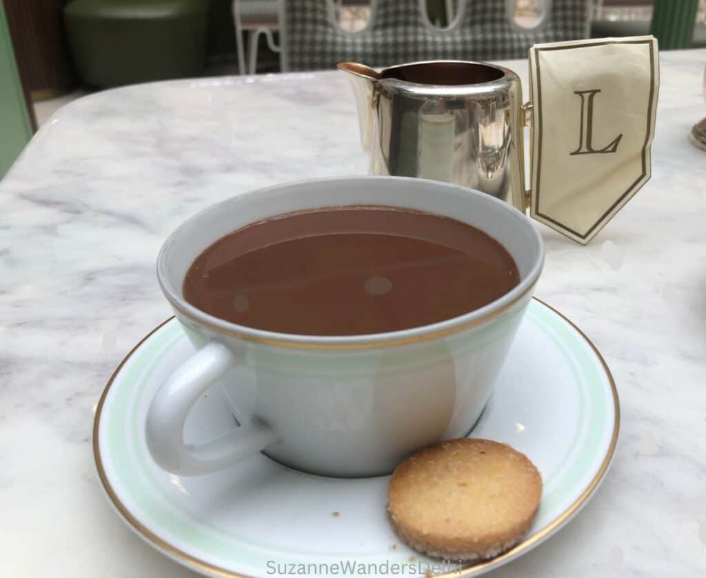 A tea cup of hot chocolate with a silver pot and cookie on the plate all on a white marble table
