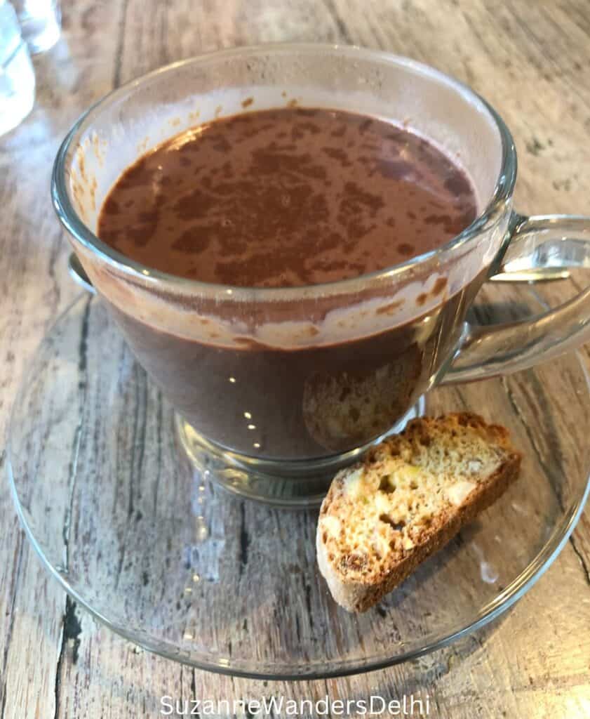 Glass cup and saucer with hot chocolate and a biscotti on a light wooden table, one of the best hot chocolates in Delhi