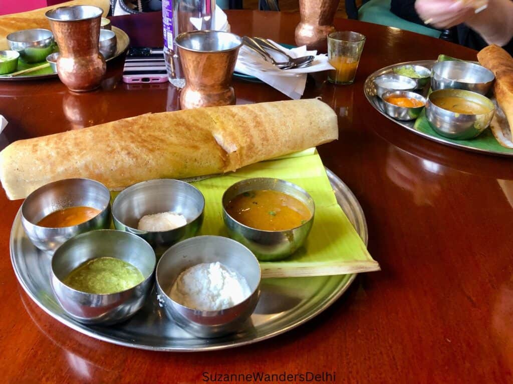 A silver plate with rolled dosa and 5 bowls of condiments on a green banana leaf - the food is one of the reasons Delhi is worth visiting