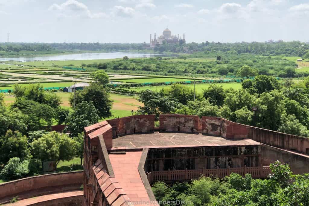 View of the Taj Mahal in the distance across green fields, a lake and part of Agra Fort