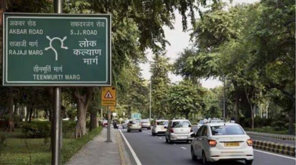 A road sign on Teenmurti Marg in Hindi and English with treed street beside and white cars