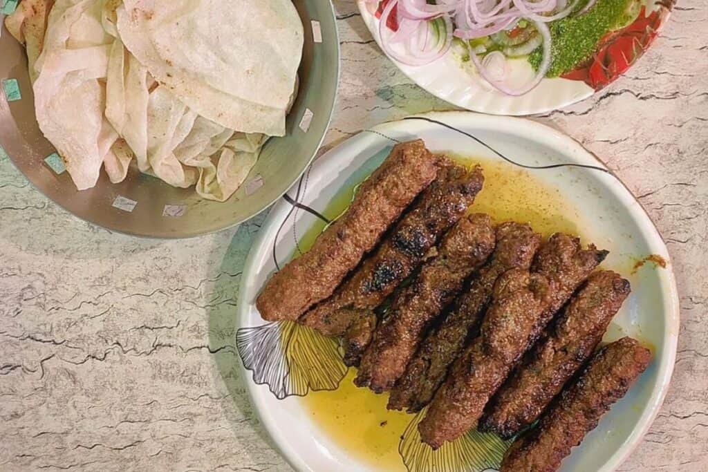 A plate of seekh kabobs in a poolof butter with a metal bowl of rumali rot on a light coloured table