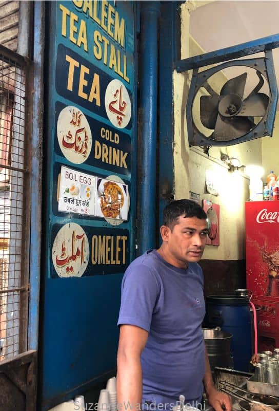 A chai seller in blue shirt standing in front of his stall with big blue sign behind him
