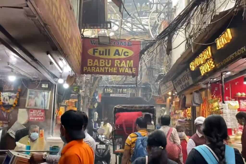 a view of Paranthe Wali Gali with parantha shop signs, pedestrians and a jumble of overhead electric wires