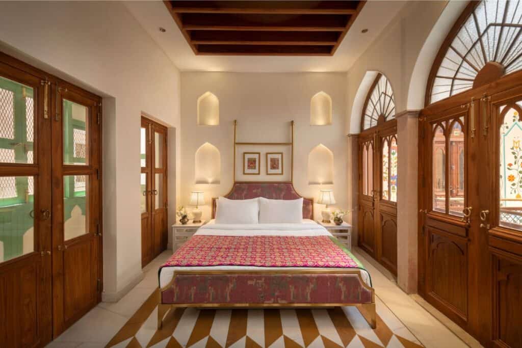 a guest room at Haveli Dharampura with a king bed int the middle and high headboard, and arched windows in teak wood