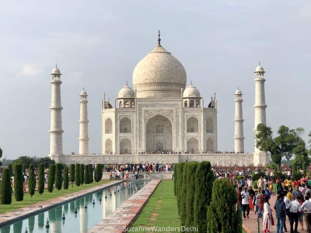 Front view of Taj Mahal in daylight, with crowds of visitors on left and long pool of water flanked by rows of small trees down the middle