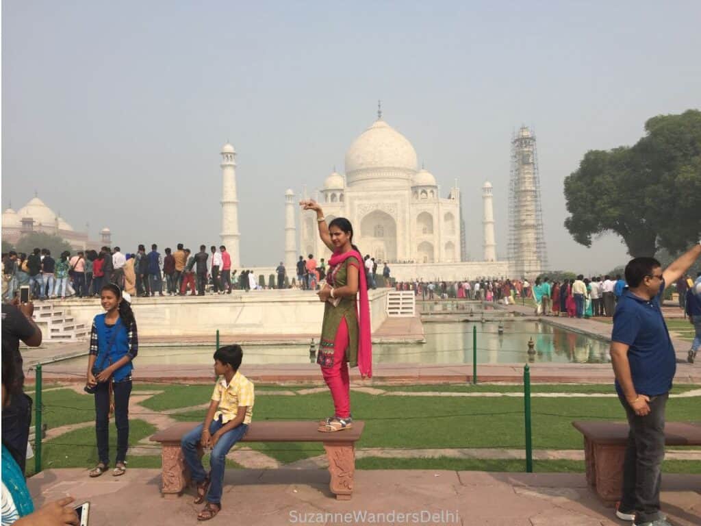 An Indian girl standing on a bench with Taj Mahal in background and many people around. The distance from Delhi to Agra is not far - you should go early to avoid the crowds like this.