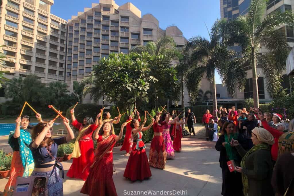 A group of international women in red saris dancing with sticks over their heads outside under palm trees