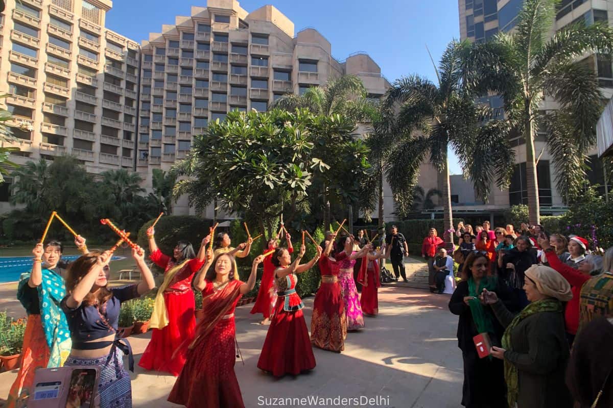 Group Asian women in red saris dancing outside holding sticks above their heads with people watching and palm trees