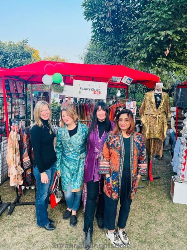 Four women standing on a lawn in front of stand with a red canopy selling clothing at the Tamana Winter Carnival in Delhi
