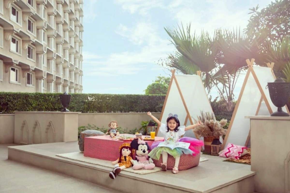 girl child sitting on round pink chair outside surrounded by stuffed animals with children's tents behind her