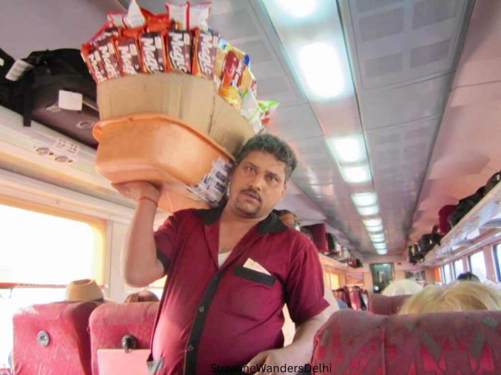 A man in red shirt walking down the aisle of a train with a big basket of chocolate bars on his shoulder