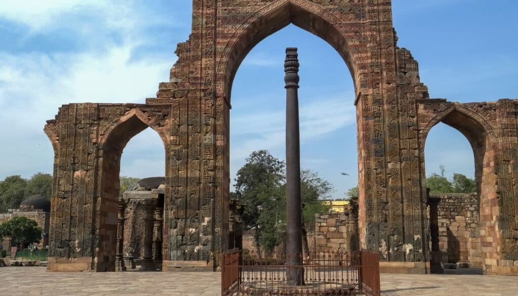 the Iron Pillar of Delhi surrounded by iron gate viewed in the middle of a large arch with blue sky