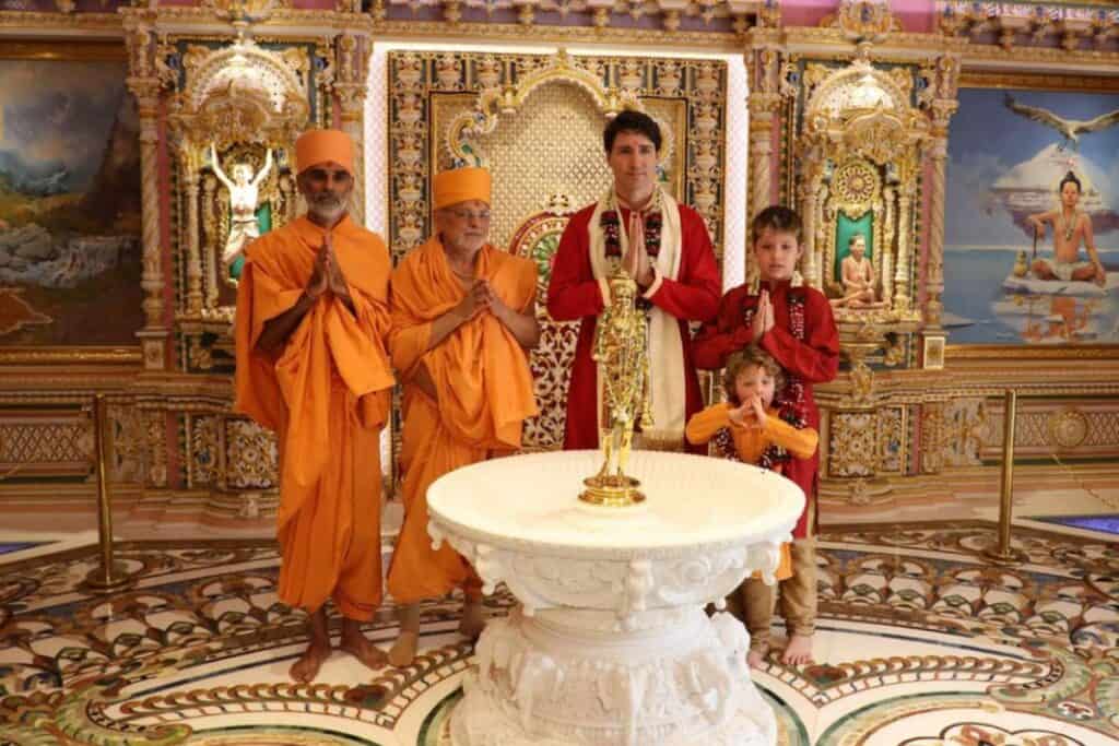 Justin Trudeau and two children in traditional Indian clothing with two orange clad yogis standing behind a small white fountain inside Akshardham Temple, Delhi