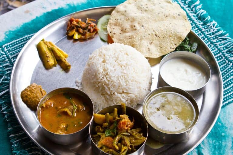 a metal thali tray with rice, papadam, pickle and 4 metal containers of food