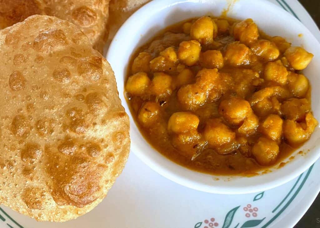 a white bowl of chana beside two puffy bhatures on a white plate, one of the most special foods of Delhi