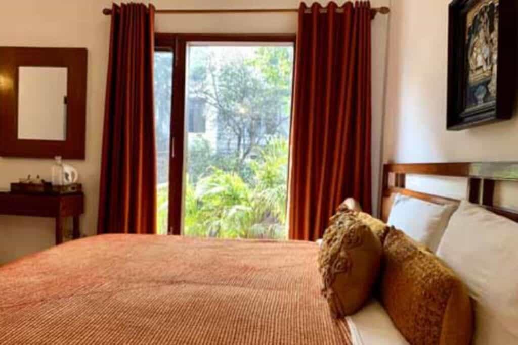 close up of large bed with burnt orange cover and large curtained window showing greenery outside at Thikana Delhi, of the best boutique hotels