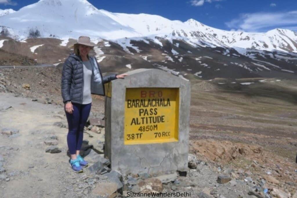 woman standing next to roadside sign for Baralachala Pass with snow peaked mountains in background