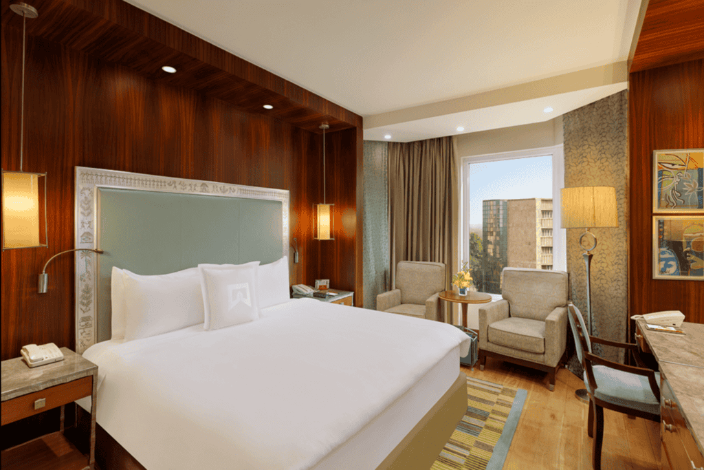 wood panelled hotel room with white king size bed and beige arm chairs at ITC Maurya in Delhi