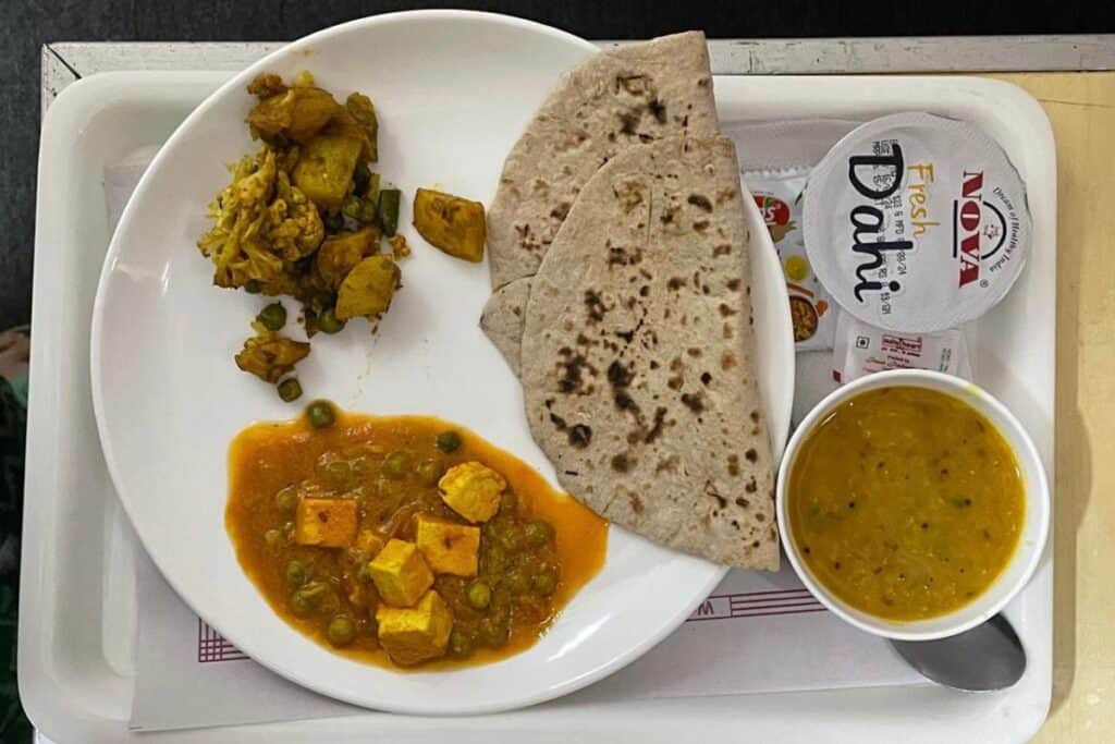 a white plate of Indian food, bowlof dal and curd on white tray on Rajdhani Express train