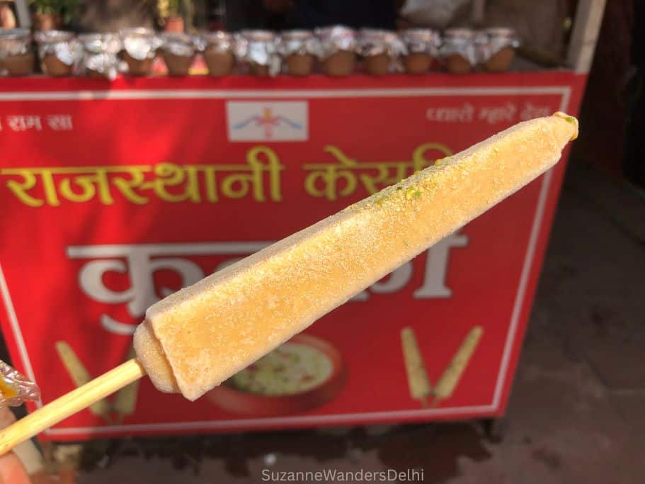 a stick of kulfi in front of a red kulfi cart in Delhi