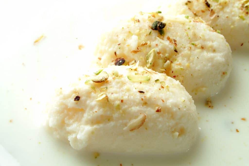 three rasmalai on a plate sitting in a shallow pool of milky syrup, one of Delhi's most popular sweets